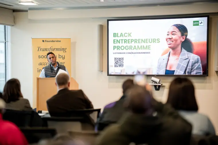 A wide view of a speaker presenting a business programme slide.