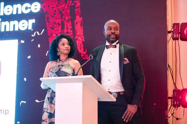 Two presenters smiling together beside the awards screen at the Black Pharma fifth anniversary gala.