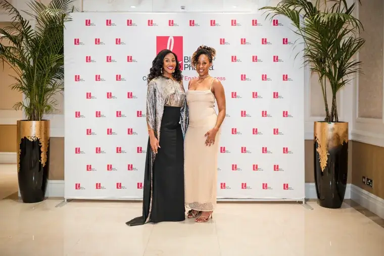Two women in evening dress posing at the photo wall at the Black Pharma fifth anniversary gala.