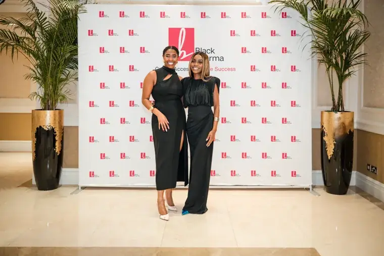 Two women posing together at the photo wall at the Black Pharma fifth anniversary gala.