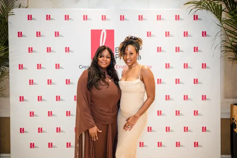 Two women smiling together in front of the branded backdrop at the Black Pharma fifth anniversary gala.